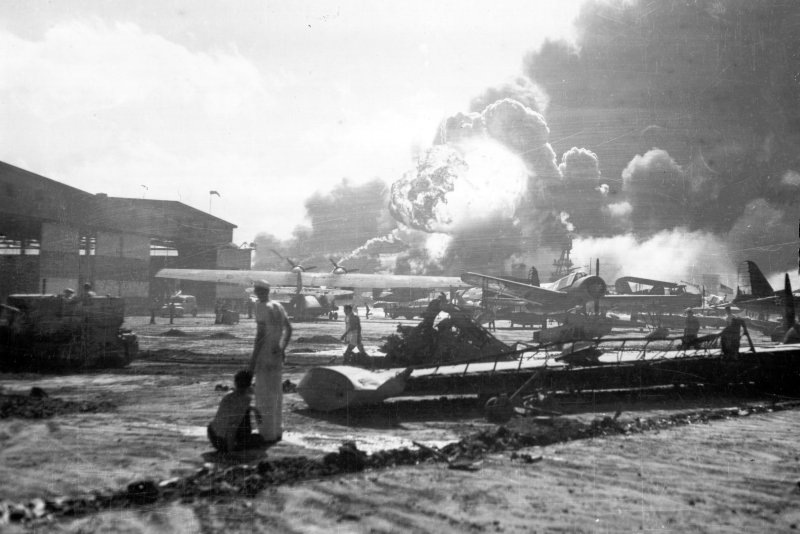 Sailors stand amid wrecked planes at the Ford Island seaplane base, watching as USS Shaw explodes in the center background December 7 1941. USS Nevada is also visible in the middle background, with her bow headed toward the left. File Photo by U.S. Navy/UPI
